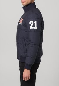 Navy blue puffer jacket with a high collar, featuring a zip closure, white number "21" on the sleeve, and an American flag emblem.