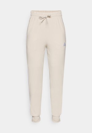 Pantalons de survêtement beige avec taille élastique, cordon de serrage et poignets fuselés. Présente un petit logo adidas violet sur la cuisse gauche. Tissu doux.