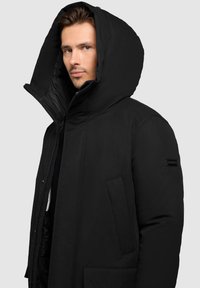 Czarna pikowana kurtka z kapturem, wysokim kołnierzem, przednim zamkiem błyskawicznym, dużymi kieszeniami, teksturowaną tkaniną i naszywką z logo na lewym rękawie.