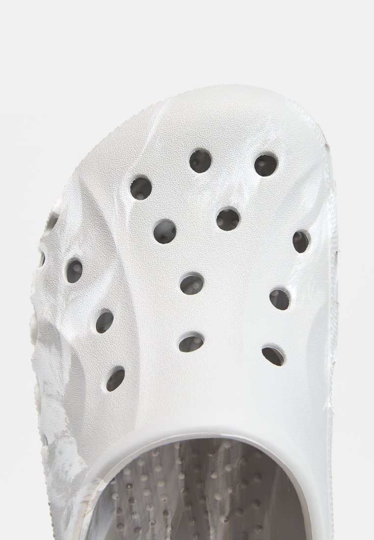 Weiße Clogs mit einer strukturierten Außenseite, die mehrere Belüftungsöffnungen und ein konturiertes Design aufweisen. Innen sind kleine erhabene Punkte für zusätzlichen Komfort.