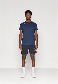 Muž stojící vzpřímeně, oblečený v tmavě modrém sportovním tričku, černých šortkách, bílých ponožkách a bílých teniskách, na jednobarevném pozadí.