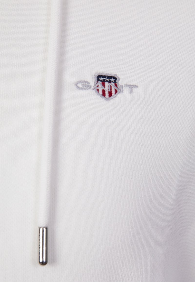 Tela blanca con un pequeño logo bordado de GANT que presenta un escudo con franjas rojas y blancas y estrellas azules, y un cordón blanco con punta metálica.