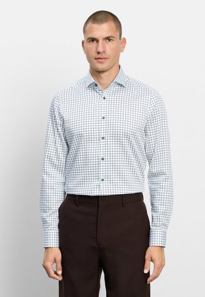 Mann trägt ein weißes langärmeliges Hemd mit blauem Kreis-Muster und dunkelbraune Hose, steht vor einem schlichten hellen Hintergrund.