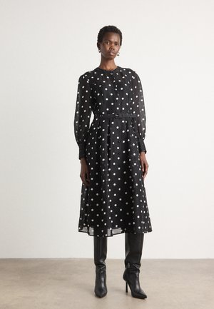 Robe noire à pois avec un devant à boutons, des manches longues, une taille ceinturée et une jupe fluide. Portée avec des bottes noires à talons hauts.