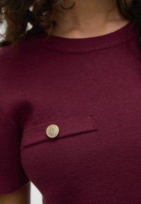 Burgundské pletené tričko s kulatým výstřihem a krátkými rukávy, se malou náprsní kapsou ozdobenou kulatým zlatým knoflíkem.