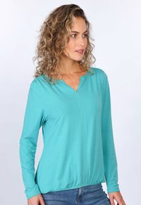 Langärmliges, türkisfarbenes Oberteil mit V-Ausschnitt und gerüschtem Saum. Hergestellt aus weichem Stoff, verfügt über eine flache Naht an der Vorderseite. Kombiniert mit blauen Jeans.