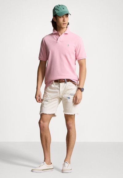 Camisa polo rosa, pantalones cortos beige desgastados, zapatillas blancas y una gorra verde. Los detalles notables incluyen un pequeño logo en la camisa y pantalones cortos deshilachados.