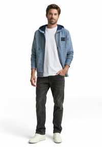 Mężczyzna w niebieskiej bluzie z kapturem, białej koszulce, czarnych dżinsach i białych trampkach, stojący z jedną ręką w kieszeni na białym tle.