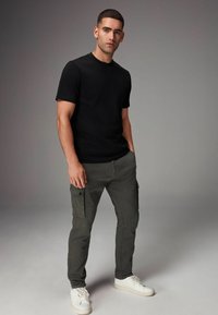 Schwarzes T-Shirt mit kurzen Ärmeln, kombiniert mit olivgrünen Cargo-Hosen mit Seitentaschen und weißen Sneakers. Einfaches, körpernahes Design.