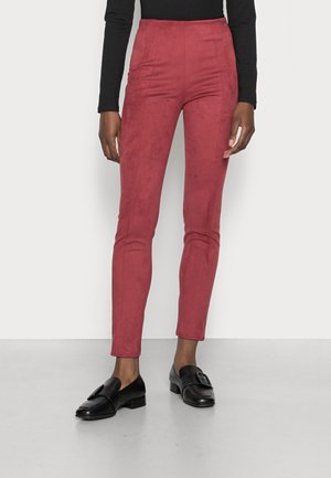 Person iført slanke roserøde bukser og sorte loafers, stående mod en ensfarvet hvid baggrund.