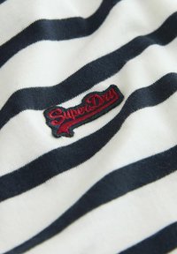Tumesinine ja valgete triipudega puuvillane kangas, millel on keskelt välja broderitud punane "SuperDry" logo. Pehme tekstuur.