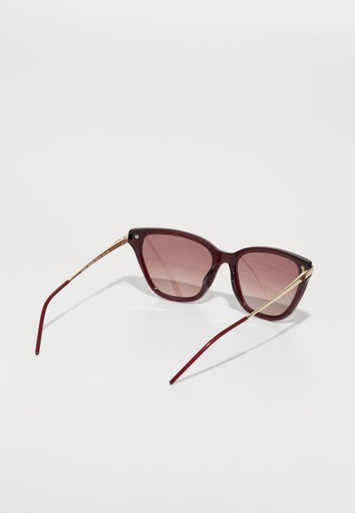 Burgundy solglasögon med gradientlinser, vinkelaktig cat-eye-form, detaljer i guldtonad metall och tunna burgundy-bågar. Strukturerad yta.