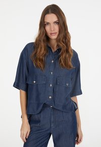 Denim shirt in dark blue, short sleeves, button-up front, two chest pockets, and visible stitching details. Smooth texture and casual design.