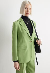 Grüne Blazer aus glattem Stoff, mit einem einzelnen Knopf, Reverskragen und seitlichen Taschen. Über einem schwarzen Rollkragenpullover und mit einer schwarzen Tasche getragen.