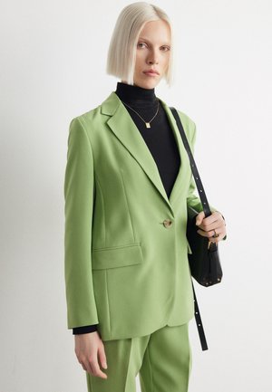 Green blazer in smooth fabric, featuring a single button, notch lapels, and side pockets. Worn over a black turtleneck with a black bag.