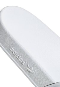 Vit lädersandal med slät yta och rund tå. Har präglad Adidas-logotyp och kontrasterande svart kant.