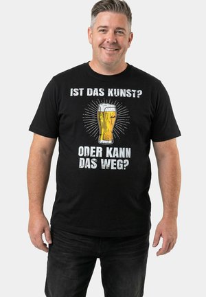Uomo sorridente, indossa una maglietta nera con un'illustrazione di un bicchiere di birra e testo in tedesco: "Ist das Kunst? Oder kann das weg?" su uno sfondo chiaro.