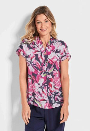 Blouse à manches courtes floral avec un fond marine, ornée de grands motifs de fleurs roses et vertes, coupe ample et col à boutons.