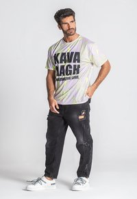 Svetlofialová grafická tričko s motívom zelených listov a tučným čiernym textom. Nosí sa s roztrhnutými čiernymi džínami a bielymi teniskami so sivými šnúrkami.