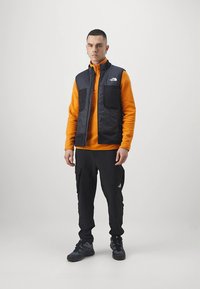 Μαύρο γιλέκο με επένδυση πάνω από πορτοκαλί φλις, συνδυασμένο με μαύρο παντελόνι cargo και σκούρα παπούτσια. Διαθέτει λογότυπο, δύο μπροστινές τσέπες και κλείσιμο με φερμουάρ.