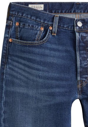 Gros plan sur l'avant d'un jean en denim bleu avec bouton, passants de ceinture et petites poches avant ornées de rivets en cuivre.