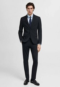 Veste de costume noire et pantalon avec une coupe ajustée, assortis à une chemise bleu clair et une cravate marine. Tissu lisse et chaussures noires élégantes.