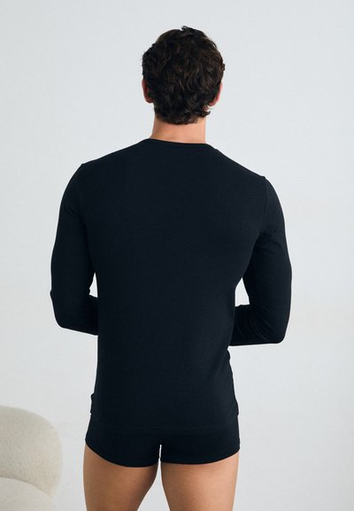 Chemise noire à manches longues en matériau lisse, design ajusté, avec encolure ronde, portée avec un short noir assorti, vue de dos.