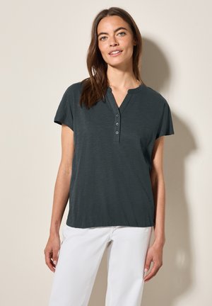 Polo shirt gris foncé à manches courtes avec une patte de boutonnage et un col en V, fabriqué en tissu doux et léger, porté avec un pantalon blanc.