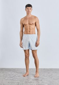 Ανοιχτό μπλε μαγιό σορτς από ελαφρύ ύφασμα με ελαστική μέση και πλευρικές σχισμές, που φοριέται από μοντέλο που στέκεται σε γκρι επιφάνεια.