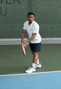 Jogador de ténis com uma t-shirt polo branca e calções pretos, segurando uma raquete e uma bola num court com rede verde ao fundo.