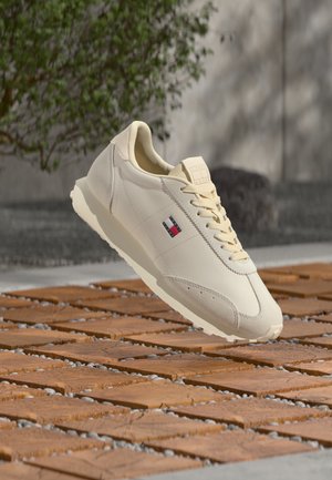 Beige casual sneaker with laces and a small red, white, and blue logo, hovering above wooden tiles with gravel in between.