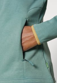 Main dans la poche d'une veste bleu sarcelle avec un poignet orné de bordure dorée et une petite étiquette en tissu vert près de l'ourlet, montrant le détail du tirette de la fermeture éclair.