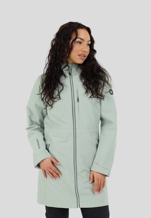 Donna con cappotto lungo verde chiaro con cerniera frontale, in piedi con le mani vicino alle tasche, guardando verso destra su uno sfondo grigio chiaro uniforme.