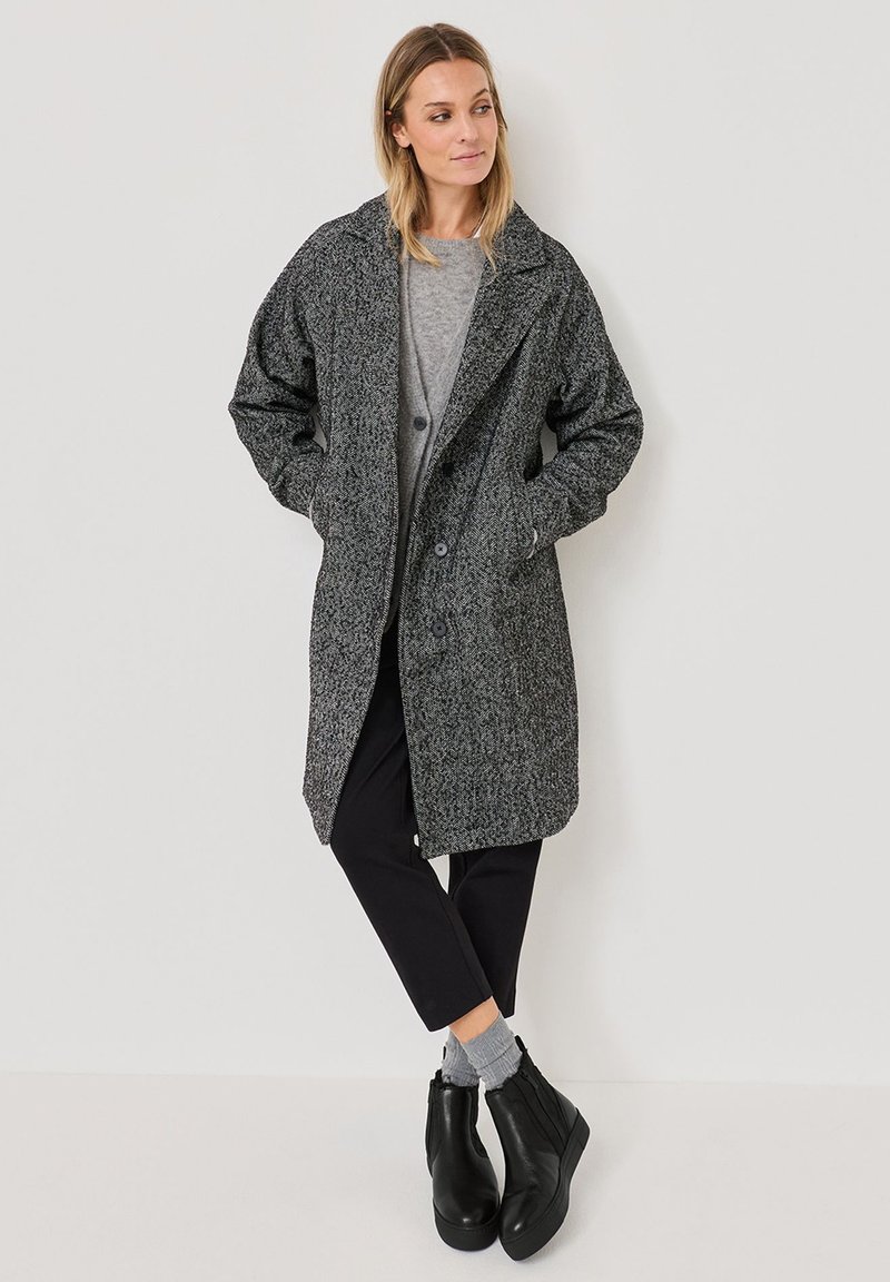 Grauer strukturierten Mantel mit Reverskragen, zwei Vordertaschen und Knopfverschluss, über einem grauen Oberteil getragen und kombiniert mit schwarzen Knöchelstiefeln.