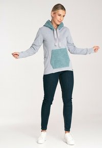 Szara bluza z kapturem i kieszenią o teksturze w kolorze teal, z sznurkiem przy kapturze i długimi rękawami, zestawiona z ciemnymi dżinsami i białymi trampkami.