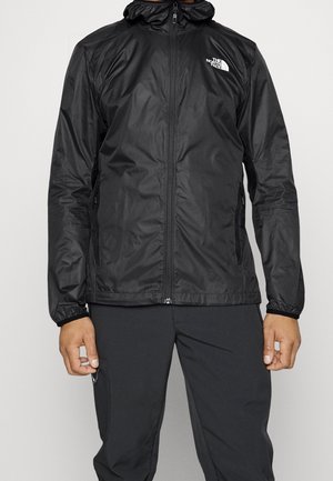 Mężczyzna w czarnej kurtce z kapturem na suwak z logo The North Face na piersi i pasujących czarnych spodniach, stojący na jednolitym tle.