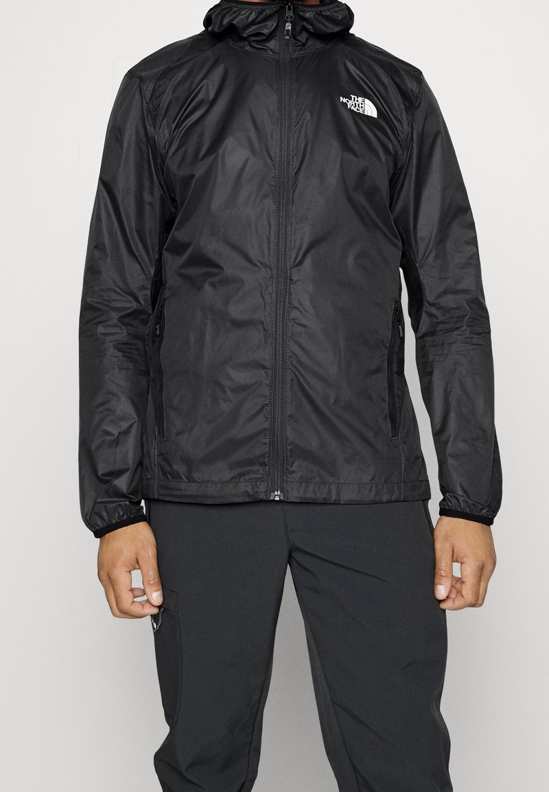Homme portant une veste noire à capuche zippée avec le logo The North Face sur la poitrine et un pantalon noir assorti, debout devant un fond uni.