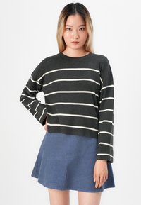 Junge Frau mit blonden Haaren, die einen dunkelgrauen Pullover mit weißen horizontalen Streifen und einen blauen Rock trägt, steht vor einem weißen Hintergrund.