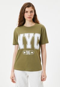 Vihreä oliivinvärinen puuvillapaita, jossa on suuri valkoinen graafinen painatus, jossa lukee "NYC" ja "96" tähtikoristeilla. Lyhyet hihat ja pyöreä kaula-aukko.