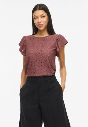 Kobieta z długimi ciemnymi włosami, ubrana w fiołkowy top z krótkim rękawem i ażurowym wzorem oraz czarne szerokie spodnie z wysokim stanem, ręce w kieszeniach.