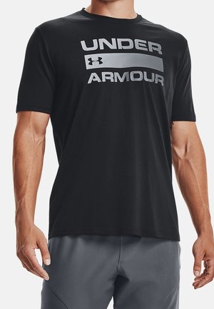 Homme portant un t-shirt Under Armour noir à manches courtes avec un logo gris et un short de sport gris, debout les bras détendus.