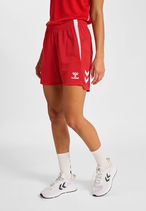 Persoon die rode sportshorts met witte strepen en witte sportschoenen met zwarte logo's draagt, staand op een lichtgrijze achtergrond.