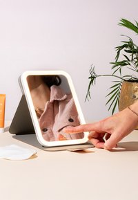 LED-beleuchteter Tischspiegel in rechteckiger Form, mit weißem Rahmen und grauem Standfuß. Eine Hand berührt die Spiegeloberfläche, die ein rosa Cordhemd reflektiert.