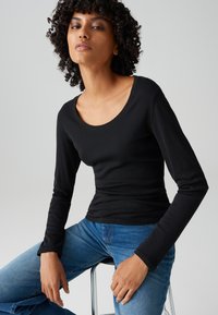 Vrouw met krullend haar, gekleed in een nauwsluitend zwart shirt met lange mouwen en een blauwe spijkerbroek, zittend op een metalen kruk tegen een effen achtergrond.