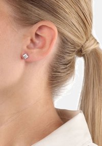 Oído derecho con un pendiente cuadrado de plata corto, pelo rubio recogido, vistiendo una camisa de color claro con cuello.