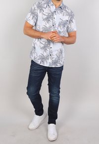 Mies, joka seisoo kädet ristissä, päällä valkoinen ja harmaa palmupainatus lyhythihainen paita, tummansiniset farkut ja valkoiset lenkkarit.