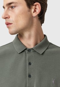 Dunkelgrünes Poloshirt aus strukturiertem Stoff, mit einem Kragen, drei Knöpfen und einem kleinen gestickten Logo auf der Brust.