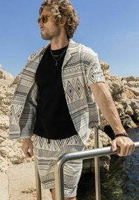 Herenoutfit met een gemusterde korte mouw overhemd en bijpassende shorts in wit en zwarte geometrische ontwerpen, gecombineerd met een zwart T-shirt met ronde hals.