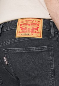 Jeans de denim negro con un parche de cuero marrón y gráficos rojos. Incluye un bolsillo trasero con una pequeña etiqueta de Levi's.