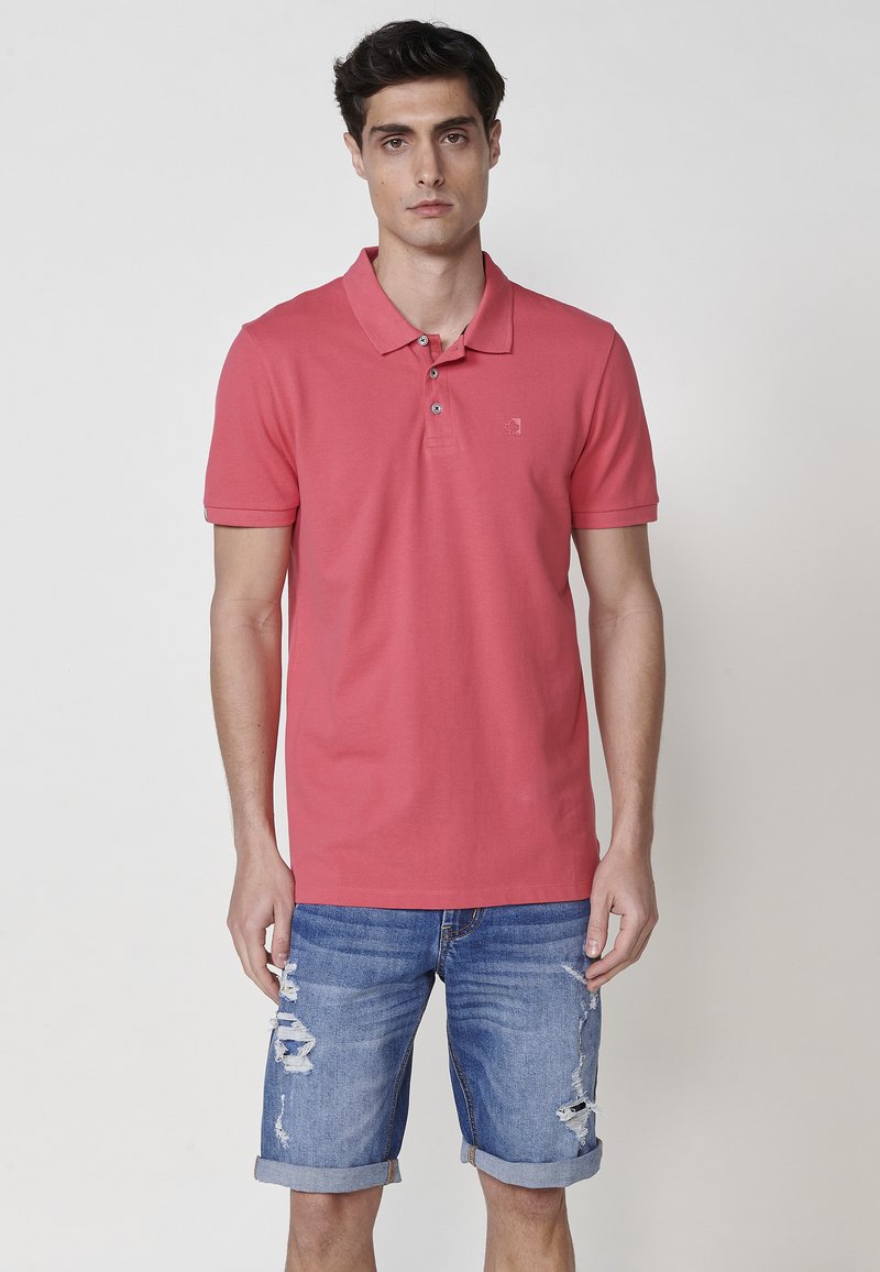 Roza polo majica s ovratnikom in tremi gumbi, v kombinaciji z raztrganimi jeans kratkimi hlačami. Majica ima subtilen logotip na prsih.